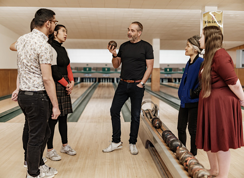 Bowling alley owner interacting with new employees. Mature man dressed in casual outfit talking to diverse group of young people. Interior of vintage bowling alley.