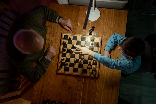 Grandfather and grandson playing chess against each other at the table in living room at home, late at night.