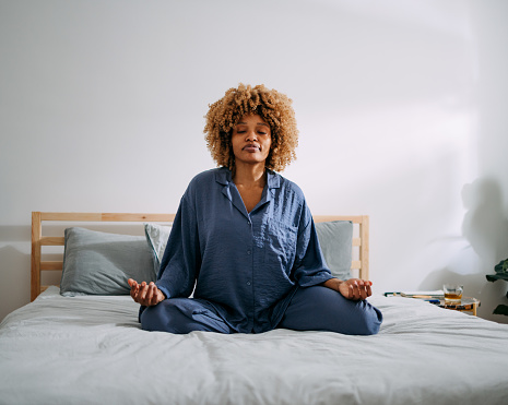Serious African American woman sitting on a bed in lotus position and meditating in her bedroom.