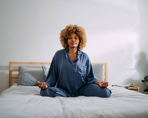 Beautiful Woman Practicing Yoga in the Morning at Home Serious African American woman sitting on a bed in lotus position and meditating in her bedroom. older woman eyes closed stock pictures, royalty-free photos & images