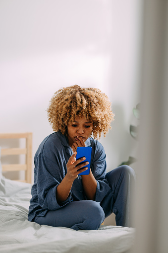 Worried African American woman in pajamas typing text message on her smartphone in the morning.