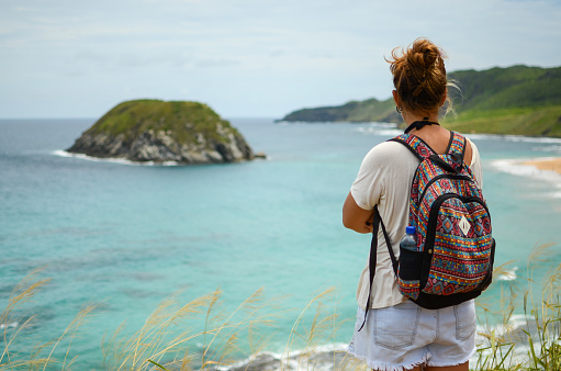 A tanned blonde girl with a backpack looking at Lion Beach, Fernando de Noronha archipelago, Brazil