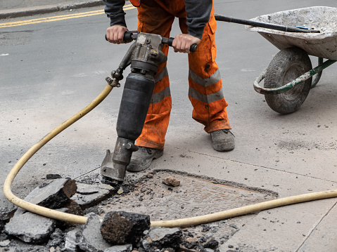 Road worker using a pneumatic drill to break up tarmac around a drain cover on a city street. The man working is wearing high visibility orange workwear and protective work boots. A wheelbarrow and spade is behind him.