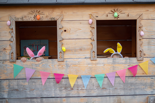 cute cheerful kids in bunny ears in the playhouse celebrate easter. brother and sister looking out the window.