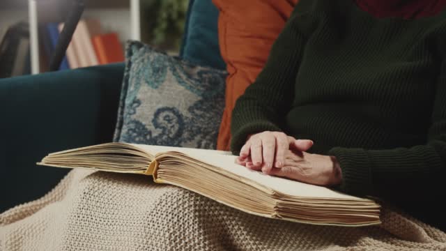 Blind woman reading braille book using fingers, sitting on sofa in living room, poorly seeing female person learning to read, home education for people with disabilities, touching letters on papers.