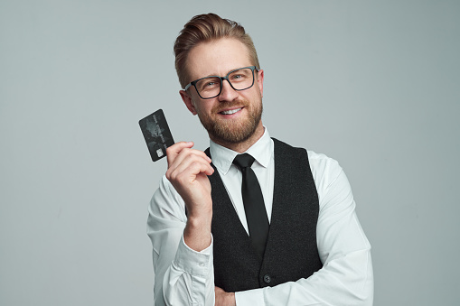 Happy male entrepreneur in glasses showing black plastic card for online purchases while looking at camera against gray background