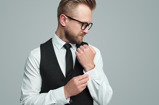 Smart male entrepreneur in elegant waistcoat and glasses buttoning sleeve of white shirt while preparing for work against gray background