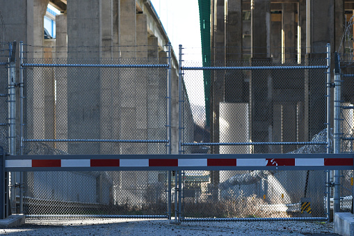 A road to an industrial complex beneath an elevated highway surrounded by barb wire with wild grass growing on it is closed and fenced off to the public.