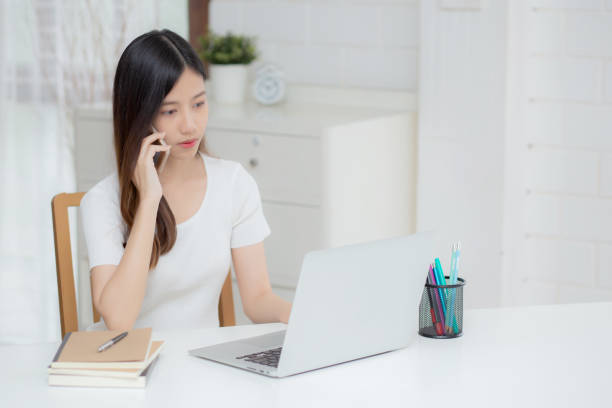 istock Young asian woman talking phone and work from home with laptop computer, freelance using notebook to internet online and speak on smartphone, domestic life, business and communication concept. 1388476906