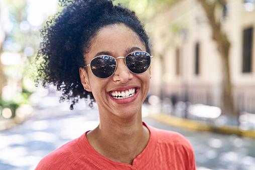 Portrait of african american adult woman wearing sunglasses looking and smiling at the camera while standing on street during daytime