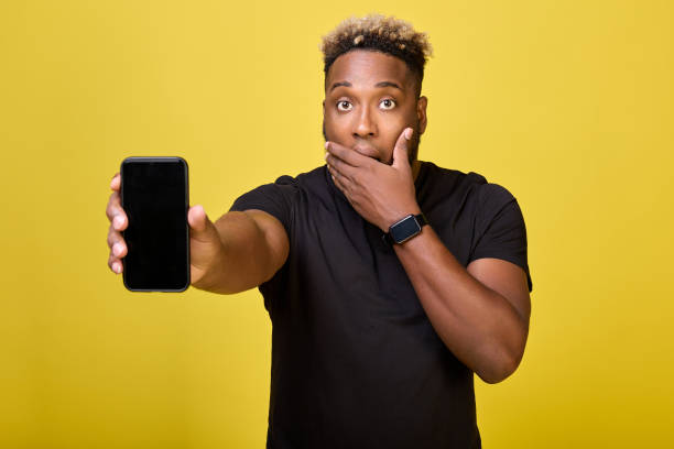 istock Surprised tall black man holds a mobile phone in his hand on yellow background 1388497991