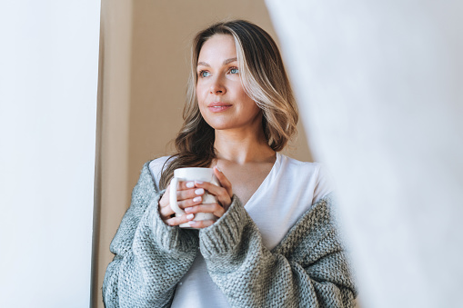 Young beautiful woman forty year with blonde long curly hair in cozy knitted grey sweater with cup of tea in hands in bright interior at home