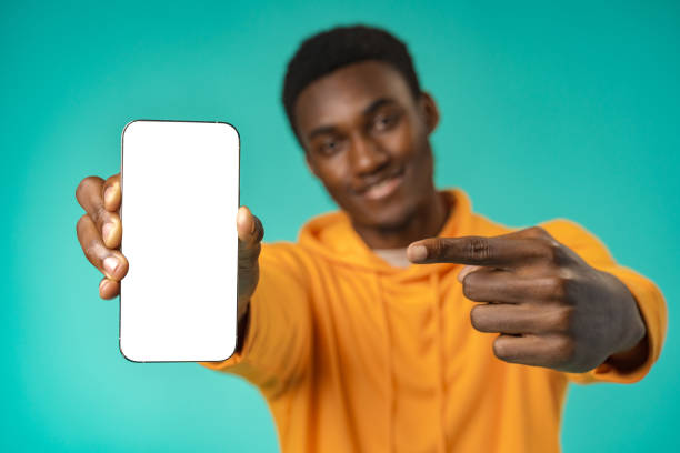 istock Smiling casual black man pointing at blank white cell phone screen in studio 1388510066