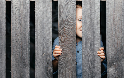 Happy smiling white boy looks out of the crack of a wooden fence. Childish curiosity. Espionage. Rural life. Child development, curiosity. The boy peered through the crack. High quality photo