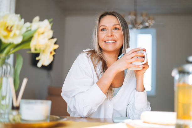 junge schöne glückliche frau mit blonden langen haaren im weißen hemd trinkt tee mit bouquet von gelben blumen in vase auf esstisch in hellem interieur zu hause - herbal tea stock-fotos und bilder