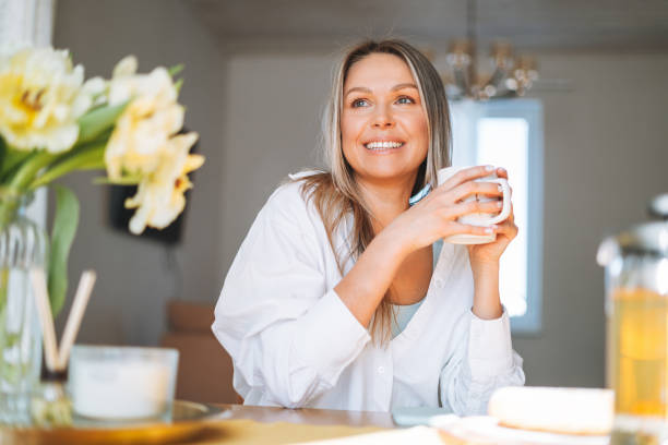 giovane bella donna felice con capelli lunghi biondi in camicia bianca che beve tè con bouquet di fiori gialli in vaso sul tavolo da pranzo in interni luminosi a casa - herbal tea foto e immagini stock