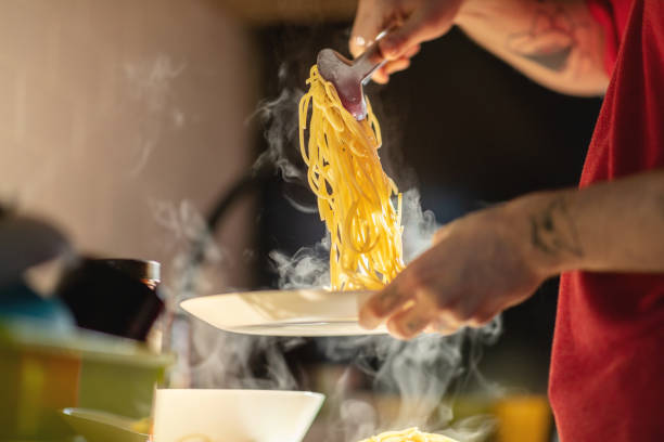 Tattooed male hand holding serving tongs and picking up steaming hot spaghetti to serve in a plate