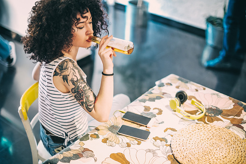 Young woman with curly hair and tattoos sitting at home and drinking non-alcoholic beer from bottle