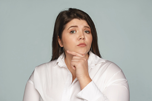 Human facial expressions, feelings and emotions. Studio image of pensive thoughtful young European businesswoman with large curvy body touching her chin, thinking over creative business ideas