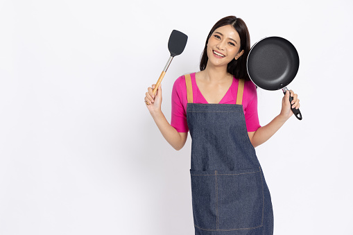 Young Asian woman housewife wearing kitchen apron cooking and holding pan and spatula isolated on white background