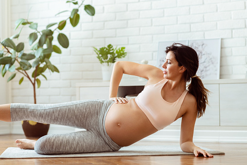 Happy pregnant woman doing yoga pilates workout and streching in the morning in her bright living room area, 30 week pregnant, side view, leg up