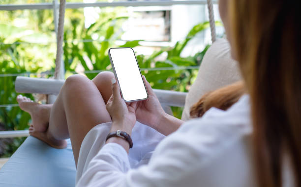 istock Mockup image of a woman holding mobile phone with blank white desktop screen while sitting and relaxing on sofa 1388530256