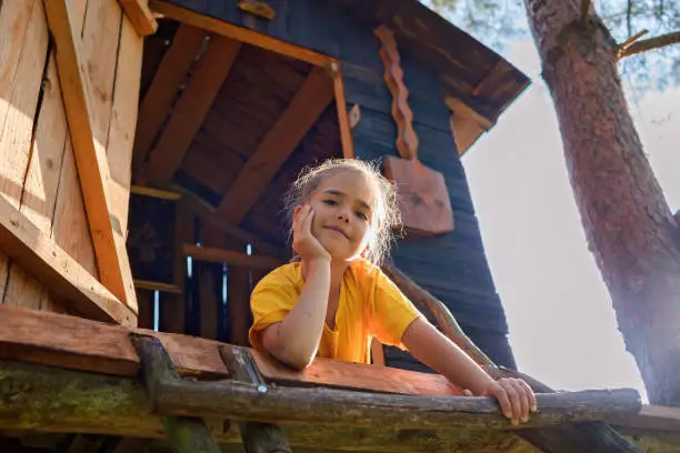 Happy pretty girl looking down from the beautiful creative handmade treehouse in backyard, summer activity, cottagecore, happy summertime in countryside, ecological outdoor playground, lower angle