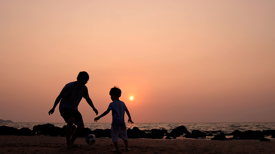 Silhouette of Asian father and son playing football or soccer on the beach at sunset, Happy family trip in summer season
