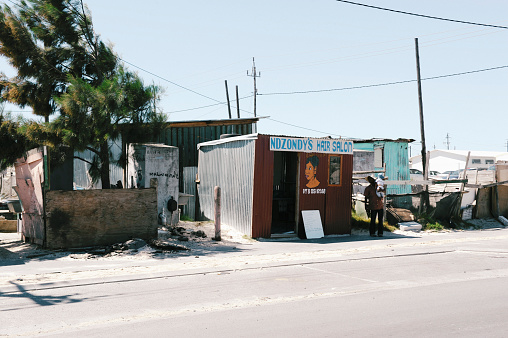 Klipheuwel, Cape Town, Western Cape, South Africa. 09 March 2016. A hair salon on the man street with one customer waiting outside.\nWith the up-coming municipal elections approaching, many people living in under-services communities like  Klipheuwel informal settlement are the forgotten people of greater Cape Town.