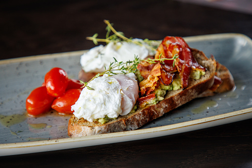 Poached egg and fried bacon slices with rosemary on top of bread slice with avocado, served with cherry tomatoes in a plate on restaurant table, gastronomic experience promotion