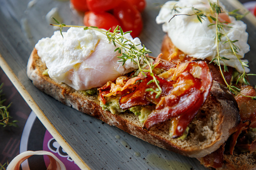 Poached egg and fried bacon slices with rosemary on top of bread slice with avocado, served with cherry tomatoes in a plate, good eating for healthy living