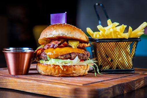 Burger in bun with bacon slices, cheese and vegetable garnish, served with French fries on pub table, gastronomic experience shared on social media