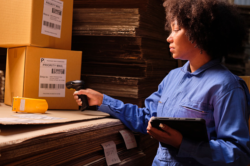 African American female worker in safety uniform using bar code scanner to check shipment orders at parcels warehouse, paper manufacture factory for the packing industry, logistic transport service.