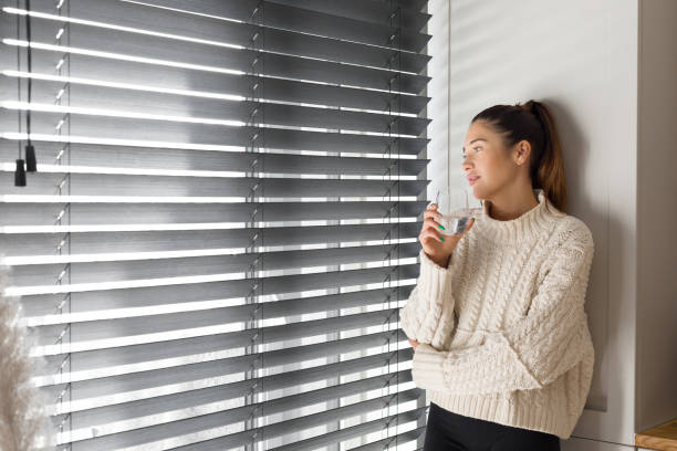 Pensive woman holding a glass of crystal clear water and looking through the blinds stock photo