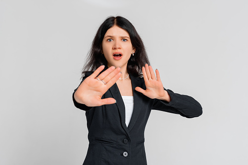 Get away from me. Scared young dark-haired girl shows hands in stop or no gesture, standing over gray background. Stay home. Social distancing. New normal behavior