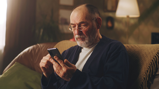 Elderly depressed man on sofa taking off glasses while holding phone with bad news and worried