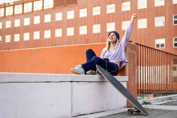 Photo of young blonde woman sitting on a wall listening music, female skater having relax in the city, happy generation z girl moving in a sustainability way of transport