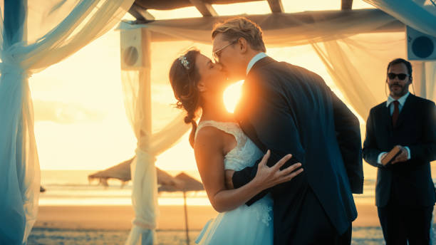 istock Beautiful Bride and Groom During an Outdoors Wedding Ceremony on an Ocean Beach at Sunset. Perfect Venue for a Couple to Marry, Exchange Rings, Kiss and Share Celebrations with Multiethnic Friends. 1388560378