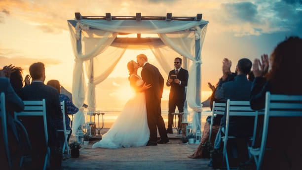 istock Beautiful Bride and Groom During an Outdoors Wedding Ceremony on an Ocean Beach at Sunset. Perfect Venue for Romantic Couple to Get Married, Exchange Rings, Kiss and Share Celebrations with Friends. 1388560559