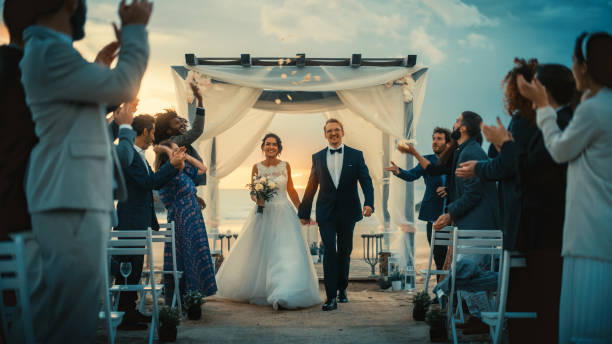 bella sposa e sposo celebrare il matrimonio all'aperto su una spiaggia vicino all'oceano al tramonto. luogo di matrimonio perfetto con i migliori amici multietnici diversi che lanciano petali di fiori sugli sposi. - wedding beach bride groom foto e immagini stock