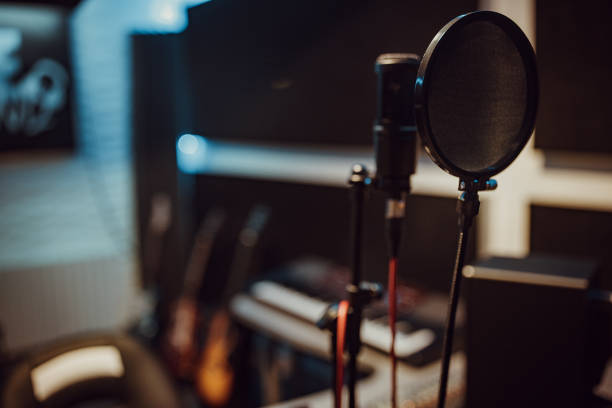 Close up of a recording microphone in a music studio. The details on the microphone can be easily seen. The microphone is silver and black with a matte metallic finish. In the back there are beautiful blue and green lights. The drums in the back reflect some shining lights.