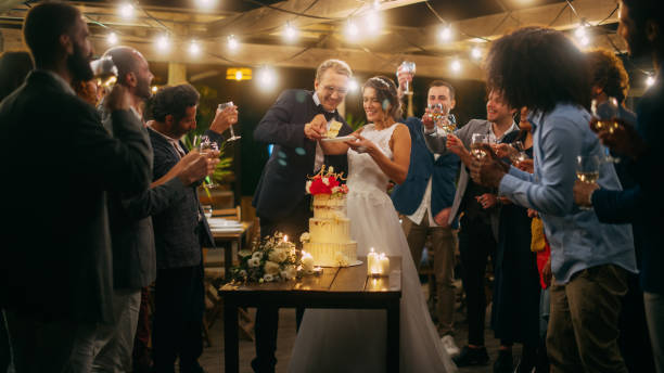 красивые жених и невеста празднуют свадьбу на вечернем приеме с многонациональными друзьями. супружеская пара стоит за обеденным столом, ц - wedding reception wine party women стоковые фото и изображения