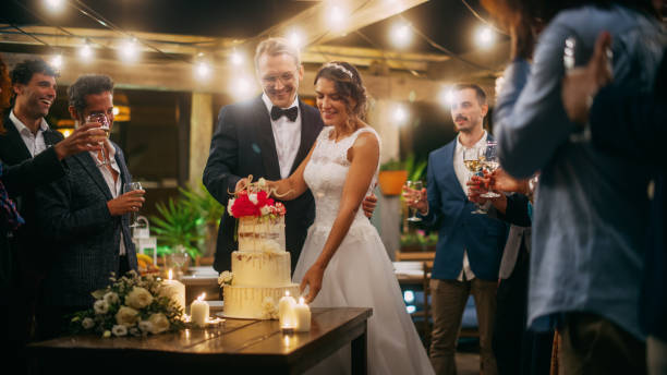 美しい新郎新婦は、多民族の友人との夜のレセプションパーティーで結婚式を祝います。ディナーテーブルに立つ夫婦、ウェディングケーキを切る。 - wedding cake newlywed wedding cake ストックフォトと画像