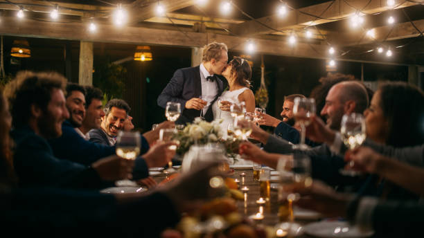 beautiful bride and groom celebrate wedding at an evening reception party. newlyweds propose a toast to happy marriage, standing at a dinner table with best multiethnic diverse friends. - wedding stockfoto's en -beelden