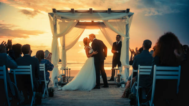 日没時のオーシャンビーチでの屋外結婚式の間に美しい新郎新婦。ロマンチックなカップルが結婚するための完璧な会場、交換リング、キスと友人とのお祝いを共有します。 - wedding newlywed bride couple ストックフォトと画像