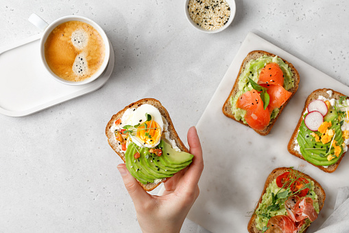 Woman hand eating a toast with avocado and egg, micro greens. Set of sandwiches with different toppings - salmon, hamon. Morning, coffee, breakfast