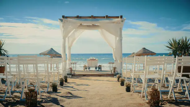 Empty Decorated Outdoors Wedding Venue with Chairs for Official Ceremony on a Beach Near the Sea or Ocean. Everything Prepared for Beautiful Romantic Marriage Celebrations. Guests are Arriving Soon.