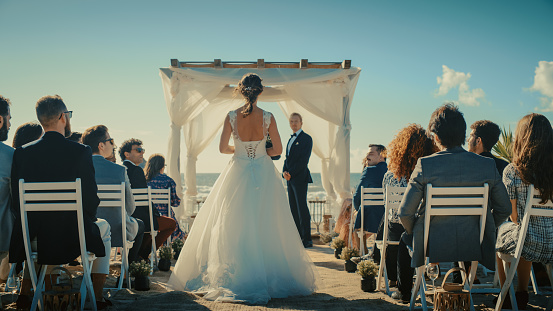 Beautiful Bride in Gorgeous White Wedding Dress Going Down the Aisle, while Groom Waits at an Outdoors Ceremony Venue Near the Sea with Happy Multiethnic and Diverse Friends.