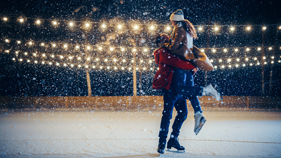 Romantic Winter Snowy Evening: Ice Skating Couple Meeton on Ice Rink and Have Fun. Pair Skating Boyfrined Lifts His Beautiful Girlfriend and Spins. Love, Dance, Embrace, Figure Skate. Wide Shot