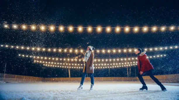 Romantic Winter Snowy Evening: Ice Skating Couple Having Fun on Ice Rink. Pair Figure Skating at Beautifully Lit Location. Boyfrined Meeting Girlfriend, Starts Dancing. Tender Young People in Love